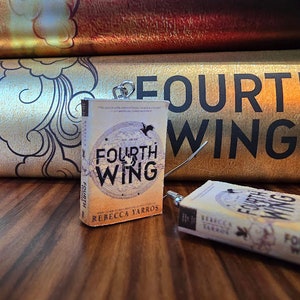 May include: Miniature book charm with the title "Fourth Wing" by Rebecca Yarros. The book has a light-colored cover with a circular design and the title in bold black letters. The background includes a gold-colored book with the title "Fourth Wing" in large black letters.