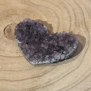 Könnte beinhalten: Ein herzförmiger Amethyst-Kristallcluster. Der Kristall hat eine tiefviolette Farbe mit einer rauen, strukturierten Oberfläche. Er liegt auf einer hellbraunen Holzoberfläche mit sichtbaren Maserungsmustern. Der Amethyst ist ein natürlicher Edelstein.