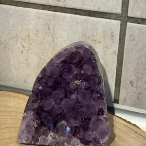 Könnte beinhalten: Eine große, rohe Amethystkristallgeode mit tiefvioletter Farbe. Der Kristall ist so geschnitten, dass ein Cluster aus funkelnden, natürlichen Amethystspitzen freigelegt wird. Die Geode liegt auf einer Holzoberfläche, mit neutralem Fliesenhintergrund.