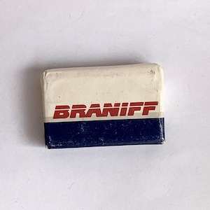 Puede incluir: Una goma de borrar rectangular con la parte superior blanca y la inferior azul marino. La palabra "BRANIFF" está impresa en rojo en el centro. Esta goma se utiliza probablemente para borrar marcas de lápiz.