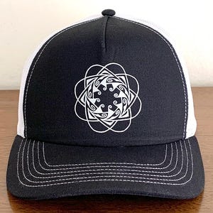 Op de afbeelding: Een zwart-witte truckercap met een wit geometrisch ontwerp aan de voorkant. De pet heeft een wit mesh-achterpaneel en een zwarte klep.