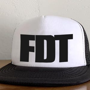 May include: A white trucker hat with a black brim and mesh back. The hat features the bold, black capital letters "FDT" across the front. The hat is sitting on a wooden surface.