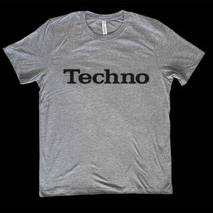 Puede incluir: Una camiseta gris jaspeado con la palabra "Techno" impresa en letras negras en el pecho. La camiseta tiene cuello redondo y mangas cortas. La tela parece suave y ligeramente texturizada.