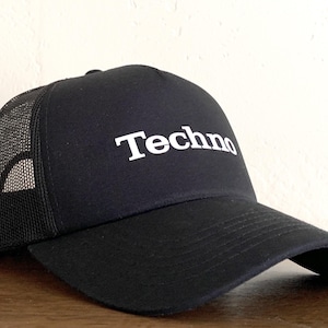 Op de afbeelding: Een zwarte truckerpet met een gestructureerd voorpaneel waarop het woord Techno in witte letters is gedrukt. De pet heeft een gebogen klep en een zwart gaaspaneel aan de achterkant voor ventilatie, wat zorgt voor een klassieke sportieve uitstraling.