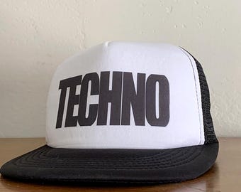 以下が含まれることがあります： 「TECHNO」の文字が黒い太字でプリントされた、白と黒のトラッカーハット。 黒いツバとメッシュのバックが付いています。