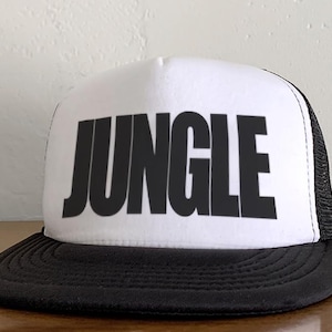 Op de afbeelding: Witte truckerpet met een zwarte klep en mesh achterkant. Het woord "JUNGLE" is in grote, vette, zwarte letters op de voorkant van de pet gedrukt.