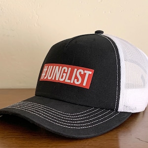 Op de afbeelding: Zwarte en witte truckercap met een rood patch waarop "#JUNGLIST" staat.