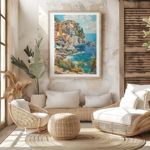 May include: A framed painting depicting a colourful seaside town built on a cliffside. The town is painted in a realistic style with a blue sky and ocean in the background. The painting is hanging on a white wall in a room with wicker furniture.