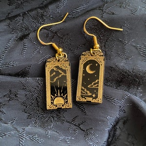 May include: A pair of gold-colored earrings with black rectangular pendants. One earring features a sun design, the other a moon and cloud design. The earrings have a decorative frame and are set against a dark, textured background.