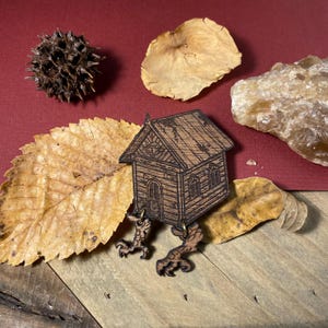 May include: A wooden house with a single eye on the front, standing on two clawed feet. The house is brown and has a weathered look. The feet are made of metal and are painted gold.