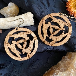 May include: A pair of wooden earrings with a circular design featuring three stylized rabbits in a circle. The earrings are made of light brown wood and have a natural finish.