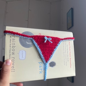 May include: A red and light blue crochet bookmark shaped like underwear, with a small bow detail. The bookmark is placed on a book titled "rainbow rowell park" and is a New York Times Bestseller.