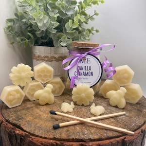 Puede incluir: Un tarro de cristal de ceras aromáticas de vainilla y canela con una cinta y etiqueta moradas. El tarro está rodeado de varias ceras de abeja con forma de flores, abejas y panales. Dos cerillas cruzadas sobre una superficie de madera.