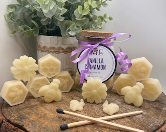 Op de afbeelding: Een glazen pot met vanille-kaneel wassmelt met een paars lint en label. De pot is omgeven door diverse bijenwas melts in de vorm van bloemen, bijen en honingraten. Twee lucifers liggen gekruist op een houten oppervlak.