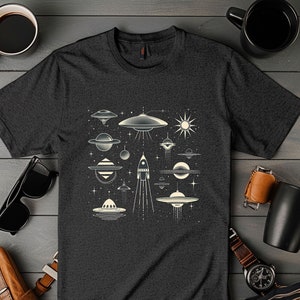 Könnte beinhalten: Ein dunkelgraues T-Shirt mit einem weißen Grafikdesign aus verschiedenen Weltraum-Illustrationen, darunter fliegende Untertassen, Planeten, Sterne und eine Rakete.