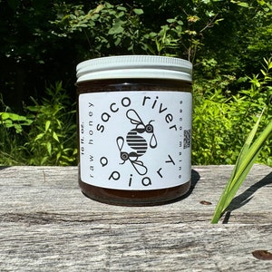 May include: A glass jar of raw honey with a white label that says "Saco River Apiary" and has a black and white illustration of two bees. The label also says "raw honey 16 fl. oz."