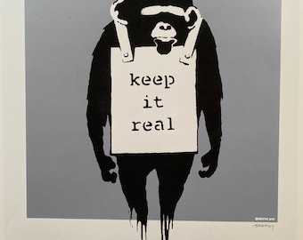 May include: A black and white print of a monkey holding a sign that says "keep it real". The monkey is wearing a black headband and the sign is attached to its neck with two straps. The background is a light gray color. The print is signed "Banksy" in the bottom right corner.