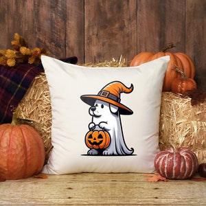 Puede incluir: Una almohada blanca con una ilustraci&oacute;n de dibujos animados de un perro con un sombrero de bruja y sosteniendo una calabaza de Halloween. La almohada est&aacute; rodeada de calabazas y balas de paja.