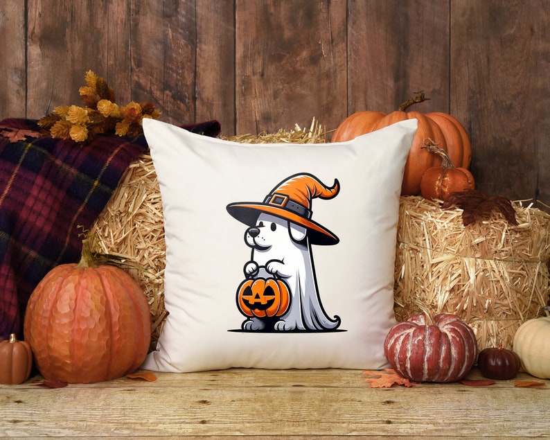 Puede incluir: Una almohada blanca con una ilustraci&oacute;n de dibujos animados de un perro con un sombrero de bruja y sosteniendo una calabaza de Halloween. La almohada est&aacute; rodeada de calabazas y balas de paja.