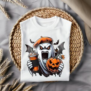 May include: White t-shirt featuring a graphic of a ghost wearing a beret, holding a pumpkin and a cup of coffee. The ghost has a scary expression with fangs and red eyes. There are bats flying around the ghost.