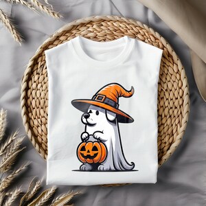 Puede incluir: Camiseta blanca con una ilustraci&oacute;n de dibujos animados de un perro con un sombrero de bruja y sosteniendo una calabaza de Halloween. El perro est&aacute; disfrazado de fantasma.