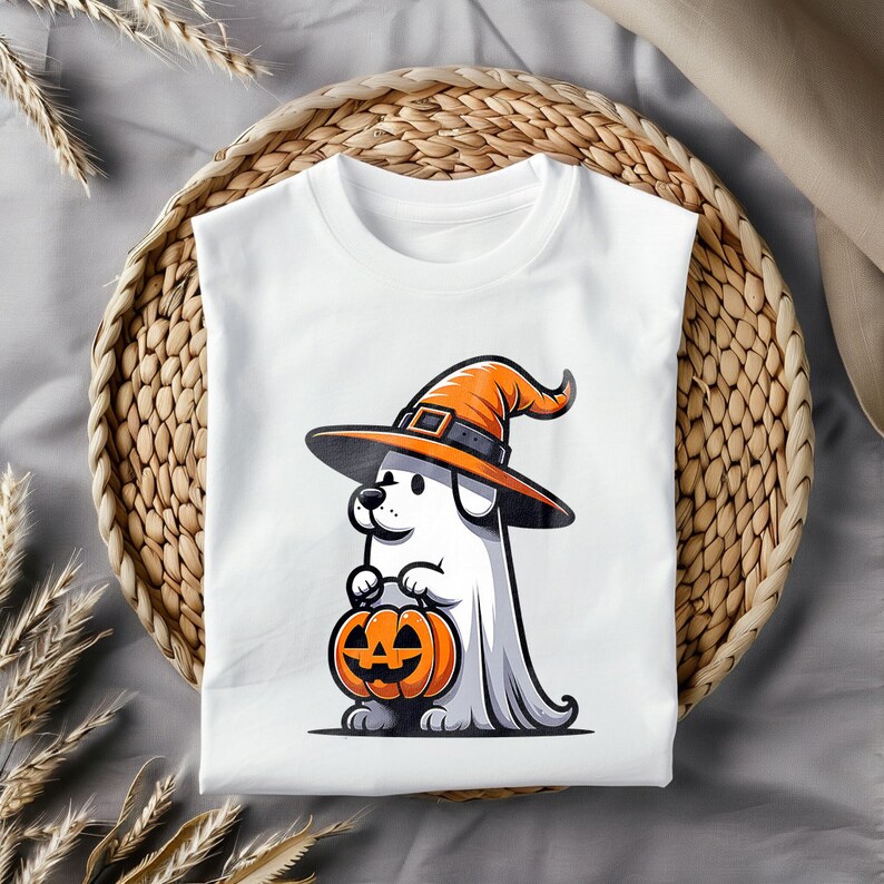 Puede incluir: Camiseta blanca con una ilustraci&oacute;n de dibujos animados de un perro con un sombrero de bruja y sosteniendo una calabaza de Halloween. El perro est&aacute; disfrazado de fantasma.