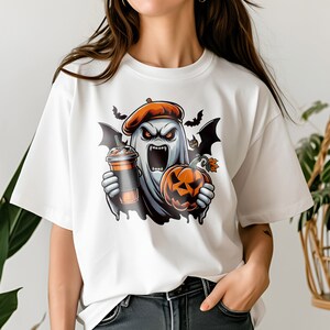 May include: White t-shirt featuring a graphic of a ghost wearing a beret, holding a pumpkin and a cup of coffee. The ghost has a spooky expression and is surrounded by bats.