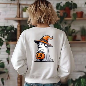 Puede incluir: Sudadera blanca con un perro fantasma de dibujos animados que lleva un sombrero de bruja y sostiene una calabaza de Halloween. El perro lleva un disfraz de fantasma blanco.