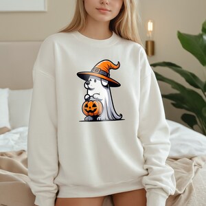 Puede incluir: Sudadera blanca con un gr&aacute;fico de un perro de dibujos animados que lleva un sombrero de bruja y sostiene una calabaza de Halloween.