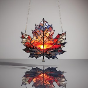 Peut inclure: Vitrail en forme de feuille d'érable avec un coucher de soleil. La feuille est composée de sections de verre rouge, orange et marron, avec une forêt et un lac en silhouette. Le soleil est un cercle jaune vif. Suspendu à deux chaînes argentées.
