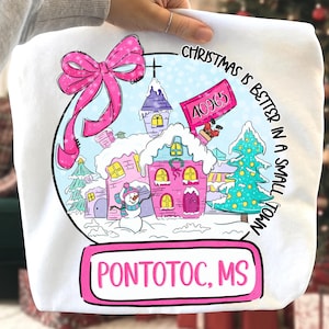 May include: White sweatshirt featuring a snow globe design with a pink bow, houses, and a snowman. The text reads "Christmas is better in a small town" and "Pontotoc, MS". A sign displays the number "40965".