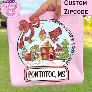 Puede incluir: Una sudadera rosa claro con un diseño de bola de nieve con temática navideña. La bola de nieve incluye un lazo rojo, casas, un muñeco de nieve y el texto "Christmas is better in a small town". También se muestra el texto "PONTOTOC, MS".