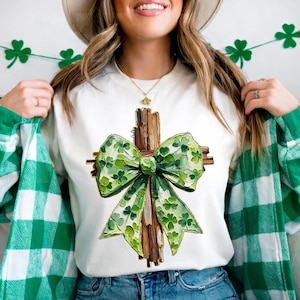 May include: White crew neck sweatshirt with a watercolour illustration of a wooden cross and a green bow decorated with shamrocks. The bow is the focal point, with varying shades of green. The sweatshirt has a St. Patrick's Day theme.