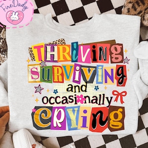 May include: A light gray sweatshirt with the words "THRIVING, SURVIVING, and occasionally CRYING" in a colorful, collage-style design. The text is set against a backdrop of various patterns and shapes, including stars and flowers.