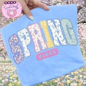 Puede incluir: Una sudadera azul claro con la palabra "SPRING" en letras grandes con estampado floral. La palabra "VIBES" está bordada en rosa debajo. La sudadera se sostiene sobre un fondo de flores.