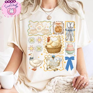 May include: Cream-colored t-shirt featuring a collection of vintage-style illustrations. The design includes a teapot, teacups, cookies, spoons, bowls, a chicken in a basket, a goose, and a shopping cart with dishes.