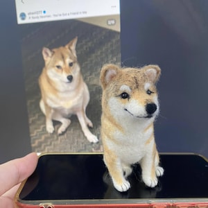 Op de afbeelding: Een naaldgevilte Shiba Inu hondje met een beige en witte vacht, zwarte ogen en een zwarte neus. Het figuurtje staat op een smartphonescherm waarop een foto van een echte Shiba Inu hond met dezelfde kleuren wordt weergegeven.