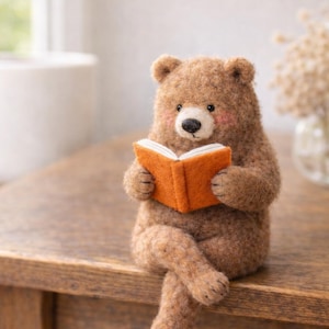 Peut inclure: Figurine d'ours en feutre marron avec des joues roses, tenant un livre orange ouvert. L'ours est assis, les jambes croisées, posé sur une surface en bois, créant une scène fantaisiste et chaleureuse.