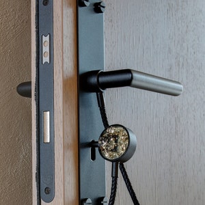 Op de afbeelding: Zwarte deurknop met een sleutelgat en een decoratieve zwarte en gouden knop. De knop is bevestigd aan een houten deur.