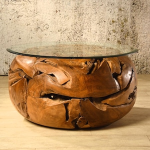 Könnte beinhalten: Ein runder Couchtisch mit einer transparenten Glasplatte. Der Sockel besteht aus aufwendig geschnitztem, naturbelassenem Holz. Das Holz hat ein raues, strukturiertes Aussehen mit verschiedenen Formen und Öffnungen. Der Tisch ist ein einzigartiges Möbelstück.