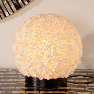 Könnte beinhalten: Eine kugelförmige Tischlampe mit floralem Design. Die Lampe besteht aus zahlreichen weißen, rosenförmigen Elementen, die ein warmes, weiches Leuchten ausstrahlen. Sie steht auf einem schwarzen Sockel und steht auf einem dekorativen Tisch. Der Durchmesser der Lampe beträgt ca. 30 cm.