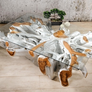 Könnte beinhalten: Ein moderner Couchtisch mit einer transparenten Glasplatte und einem einzigartigen Untergestell aus einer natürlichen, verwitterten Holzmaserung. Das Holzgestell ist hellgrau lackiert mit natürlichen braunen Holzakzenten. Ein kleiner Bonsai steht auf dem Tisch.