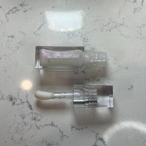 May include: A clear lip gloss tube with a rectangular cap and applicator. The lip gloss is pearlescent and the text "BELAC COSMETICS" is printed on the tube. The applicator is a white, fluffy wand.