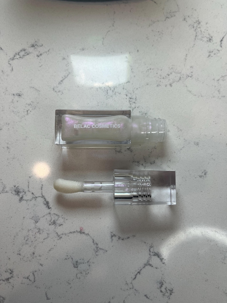 May include: A clear lip gloss tube with a rectangular cap and applicator. The lip gloss is pearlescent and the text "BELAC COSMETICS" is printed on the tube. The applicator is a white, fluffy wand.
