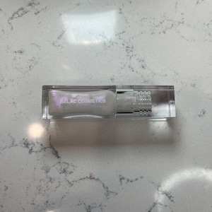 May include: A clear rectangular cosmetic tube with a silver cap and the text "BELAC COSMETICS" in silver. The tube contains a pearlescent liquid. The product is displayed on a white and gray marble surface.