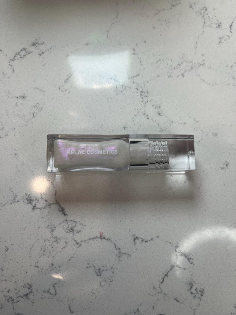 May include: A clear rectangular cosmetic tube with a silver cap and the text "BELAC COSMETICS" in silver. The tube contains a pearlescent liquid. The product is displayed on a white and gray marble surface.