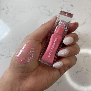 May include: A clear lip gloss tube with a pink tint and the text "BELAC COSMETICS" is held in a hand. A swatch of the gloss is visible on the hand. The gloss has a shiny, wet appearance. The tube has a clear cap.