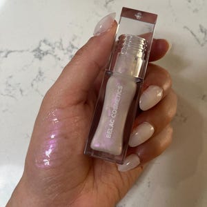 May include: A clear lip gloss tube with a pearlescent, iridescent finish. The gloss is in a clear rectangular container with the words "BELAC COSMETICS" printed on the side. The gloss is swatched on the hand, showing a pink and purple shimmer.