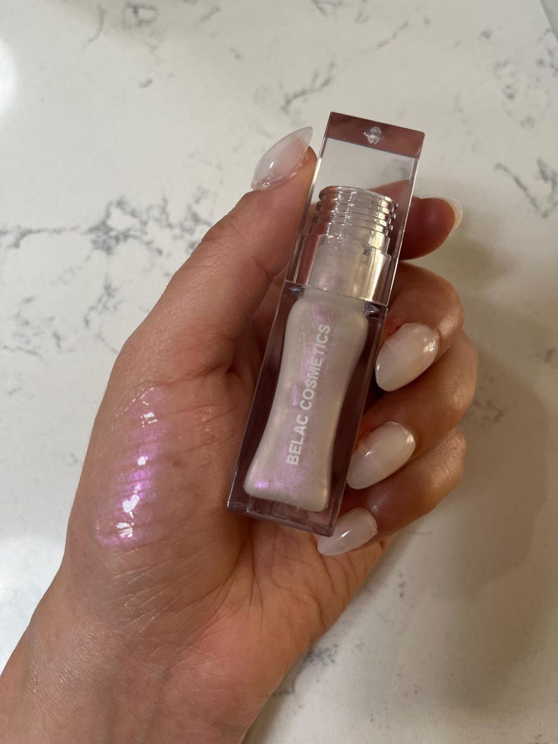 May include: A clear lip gloss tube with a pearlescent, iridescent finish. The gloss is in a clear rectangular container with the words "BELAC COSMETICS" printed on the side. The gloss is swatched on the hand, showing a pink and purple shimmer.