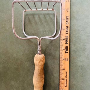 May include: A vintage potato masher with a wooden handle and a metal mashing head. The handle is light brown and the mashing head is made of silver-colored metal. A wooden ruler is next to the masher. The ruler is marked with the text "Twin Pines Farm Dairy".